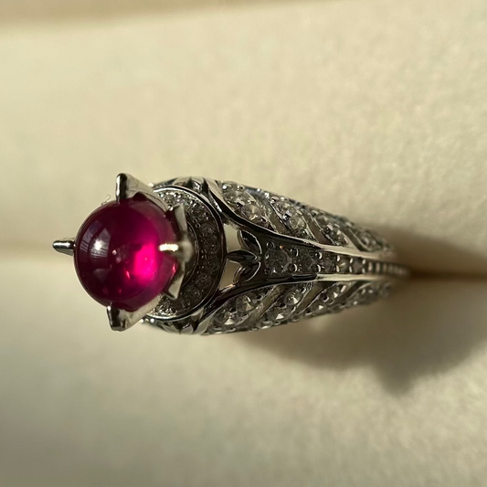 A vintage-style silver ring featuring a round, vibrant magenta-pink ruby in a prong setting, surrounded by small clear stones and an ornate filigree band, resting on a textured beige surface.