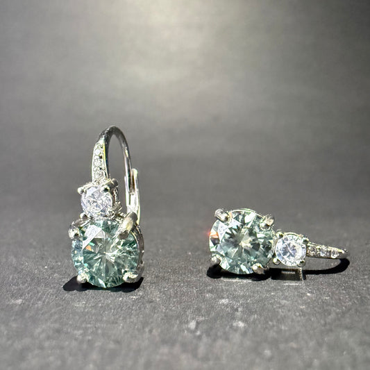 A pair of silver leverback earrings with large round-cut light blue-green moissanite and smaller clear stones.