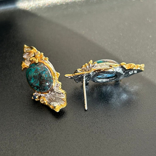 A pair of vintage-style statement earrings featuring oval dark blue-green Chrysocolla stone centers surrounded by silver metal, with textured gold leaf or wing-like side details and small dark blue stones, shown on a plain light background.