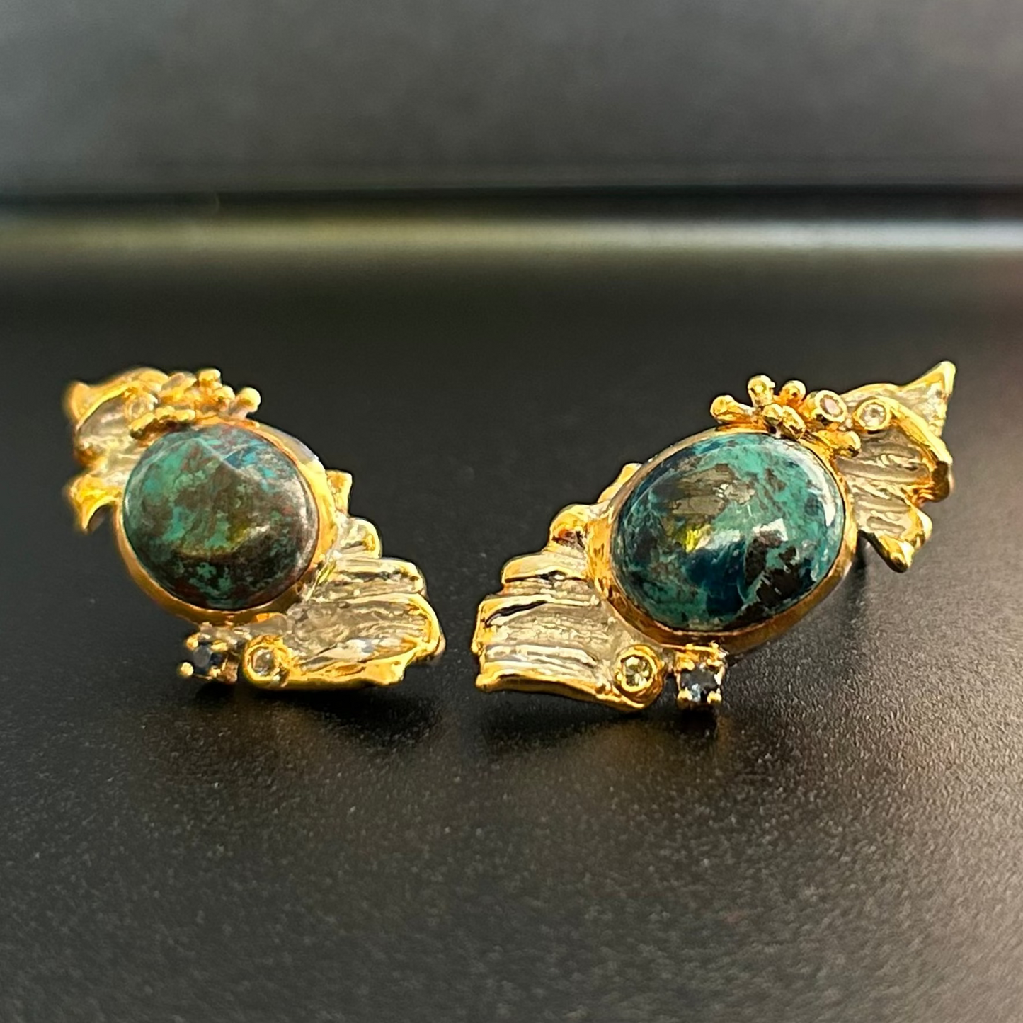 A pair of vintage-style statement earrings featuring oval dark blue-green Chrysocolla stone centers surrounded by silver metal, with textured gold leaf or wing-like side details and small dark blue stones, shown on a plain dark surface.