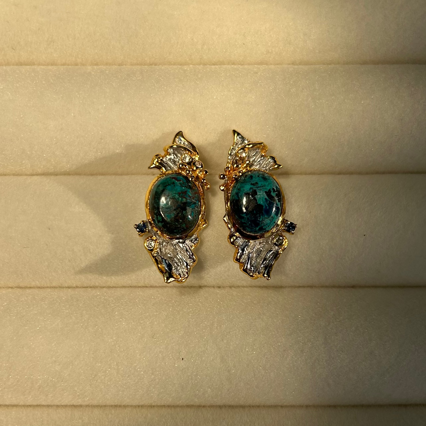 A pair of vintage-style statement earrings featuring oval dark blue-green Chrysocolla stone centers surrounded by silver metal, with textured gold leaf or wing-like side details and small dark blue stones, shown on a plain light background.