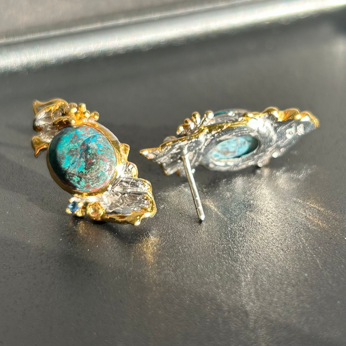 A pair of vintage-style statement earrings featuring oval dark blue-green Chrysocolla stone centers surrounded by silver metal, with textured gold leaf or wing-like side details and small dark blue stones, shown on a plain light background.