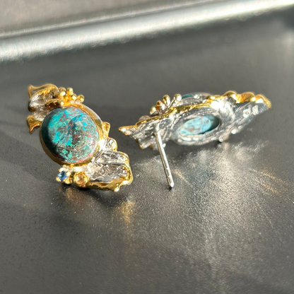 A pair of vintage-style statement earrings featuring oval dark blue-green Chrysocolla stone centers surrounded by silver metal, with textured gold leaf or wing-like side details and small dark blue stones, shown on a plain light background.