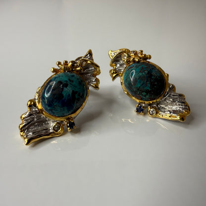 A pair of vintage-style statement earrings featuring oval dark blue-green Chrysocolla stone centers surrounded by silver metal, with textured gold leaf or wing-like side details and small dark blue stones, shown on a plain light background.