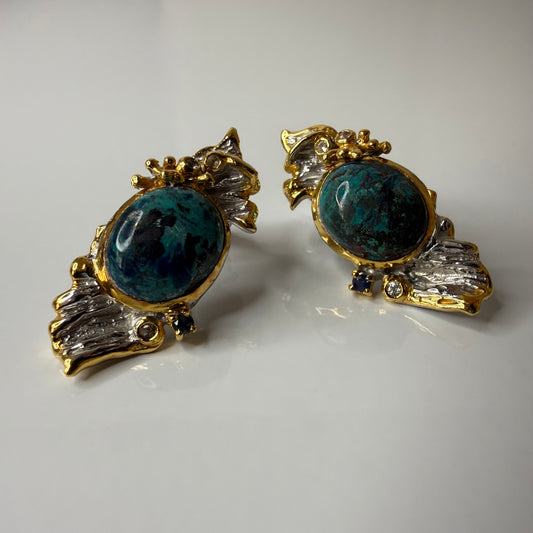 A pair of vintage-style statement earrings featuring oval dark blue-green Chrysocolla stone centers surrounded by silver metal, with textured gold leaf or wing-like side details and small dark blue stones, shown on a plain light background.
