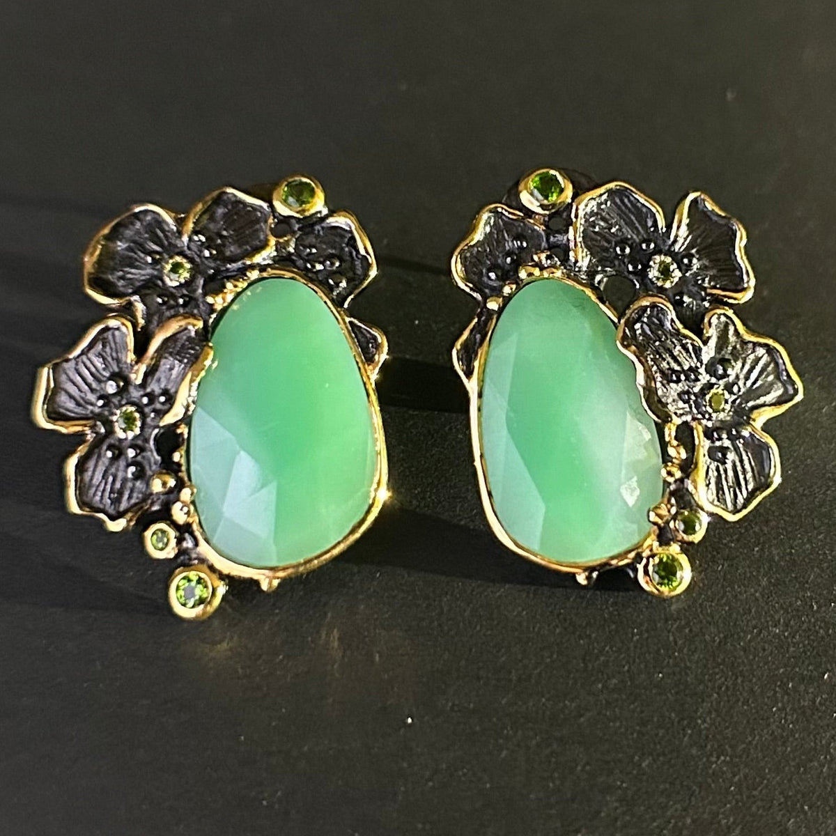 Pair of earrings with green teardrop stones and gold floral designs on a black background