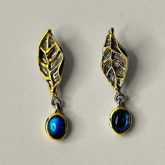 Pair of gold and silver leaf-shaped earrings with blue stones on a light gray background