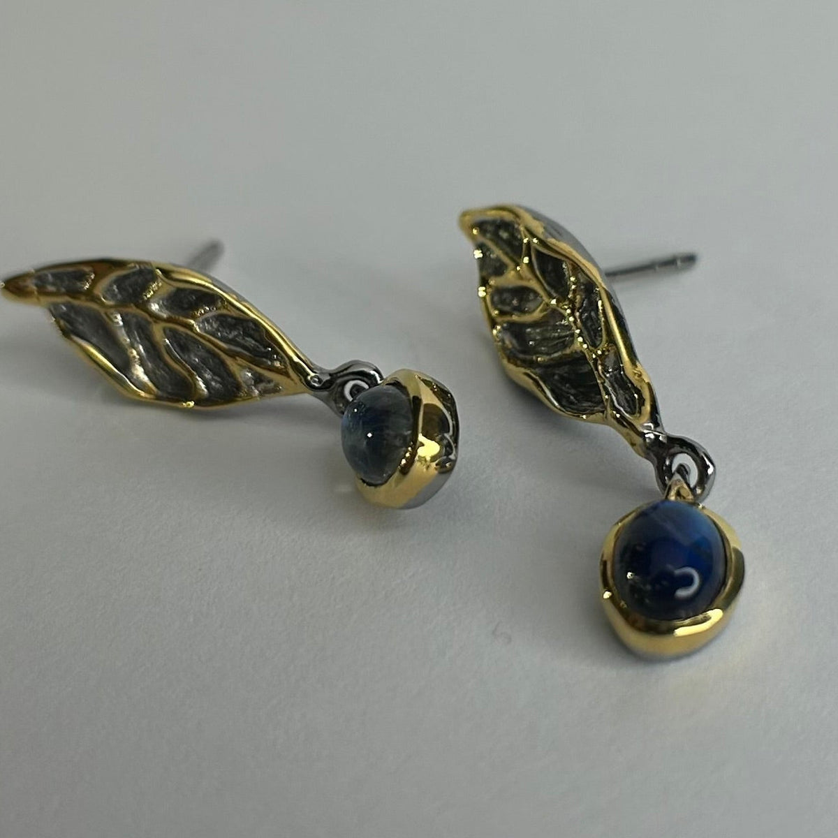 Gold and silver leaf-shaped earrings with blue stones on a light gray background