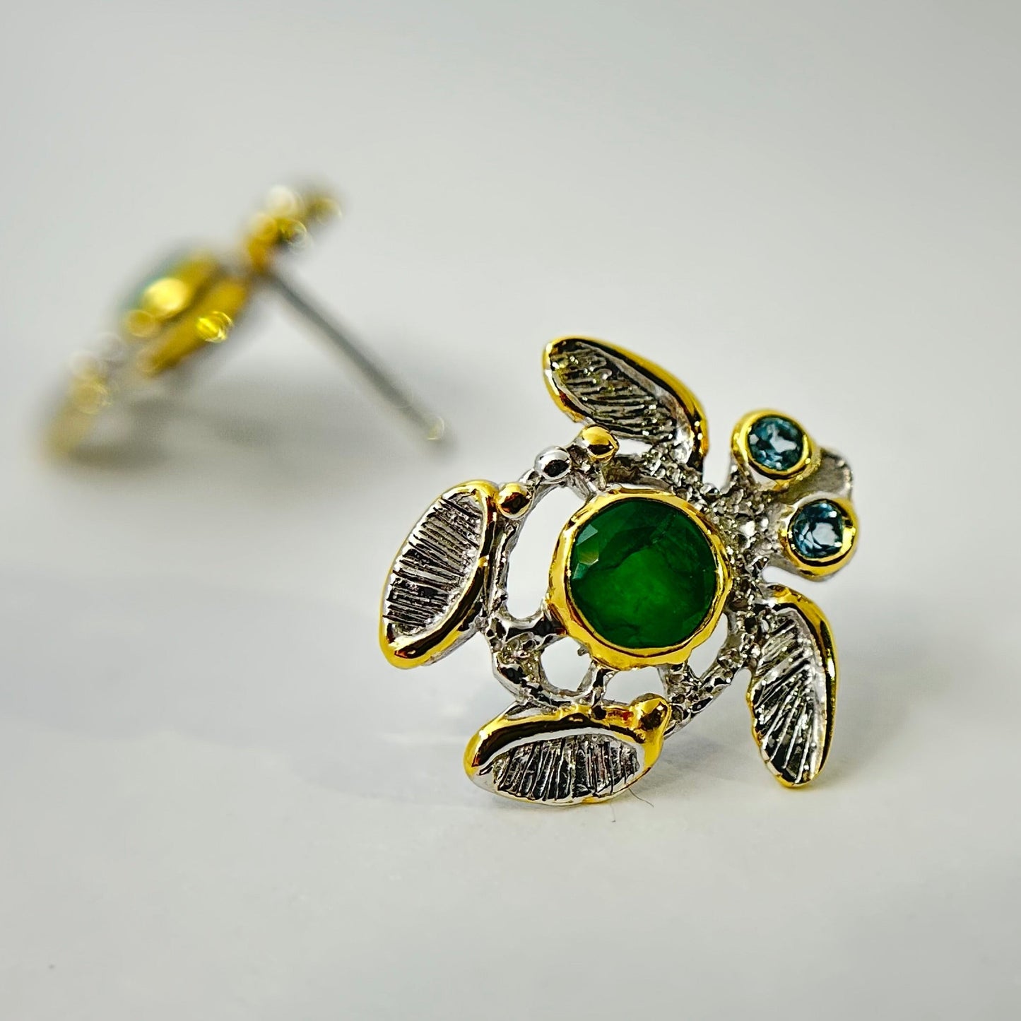 Turtle-shaped silver earrings with green emerald gemstones on a light gray background