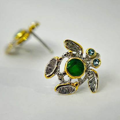 Turtle-shaped silver earrings with green emerald gemstones on a light gray background