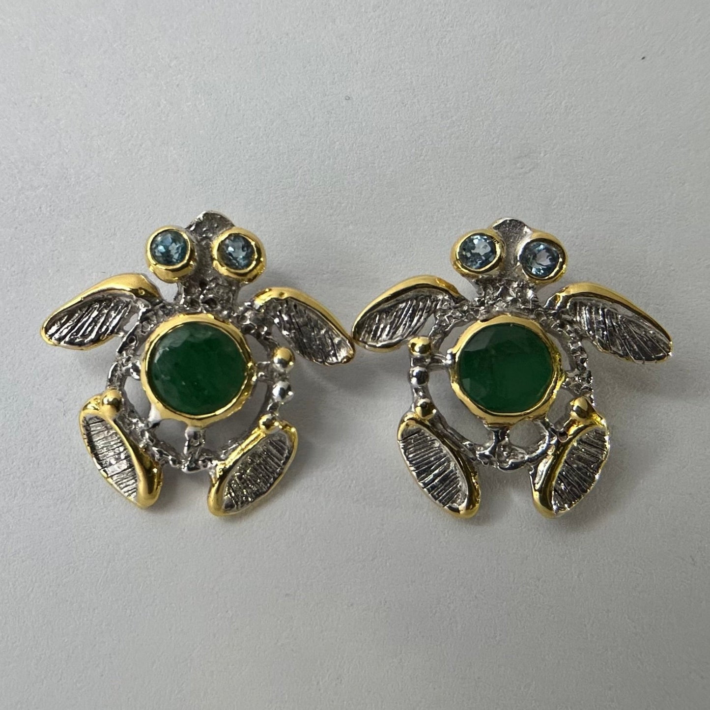 Turtle-shaped silver earrings with green emerald gemstones on a light gray background