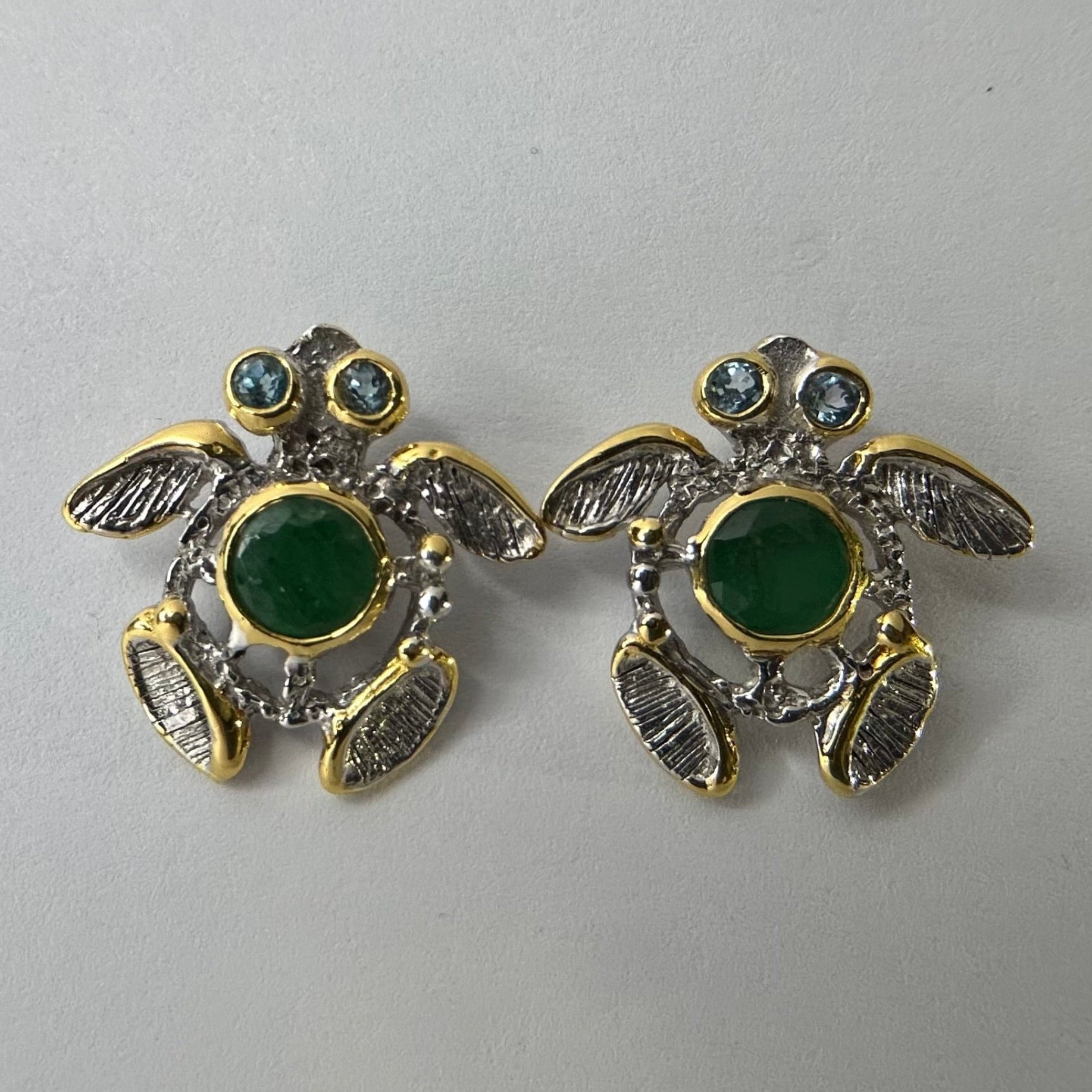 Turtle-shaped silver earrings with green emerald gemstones on a light gray background