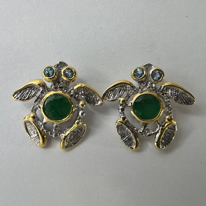 Turtle-shaped silver earrings with green emerald gemstones on a light gray background