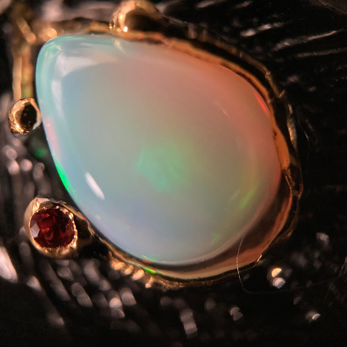 Opal gemstone in a gold setting with a red gemstone on a textured black background