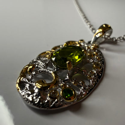 An ornate oval pendant necklace featuring a large green Peridot gemstone at the center, surrounded by textured silver filigree and small gold accents, with a matching silver chain visible against a light gray background.