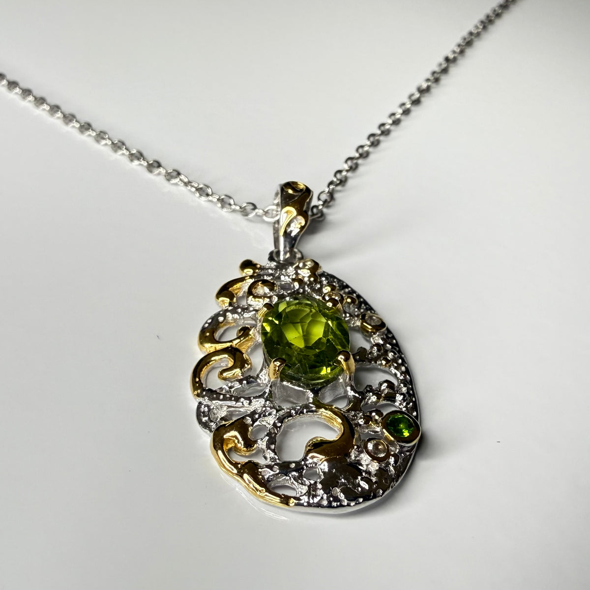 A two-tone silver and gold filigree pendant necklace featuring a central green peridot gemstone and a smaller green stone, displayed on a clean white background.