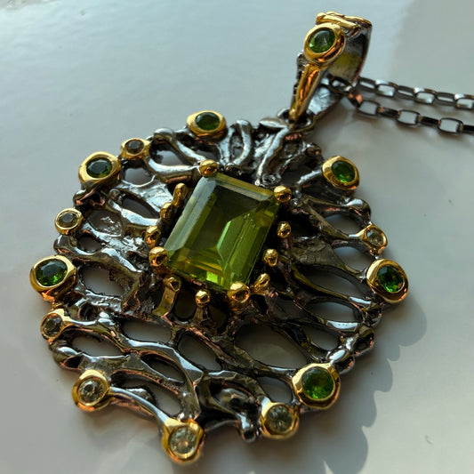 An ornate pendant necklace featuring a central rectangular green peridot gemstone surrounded by a filigree-style silver and gold setting with small green accent stones, shown on a white background.