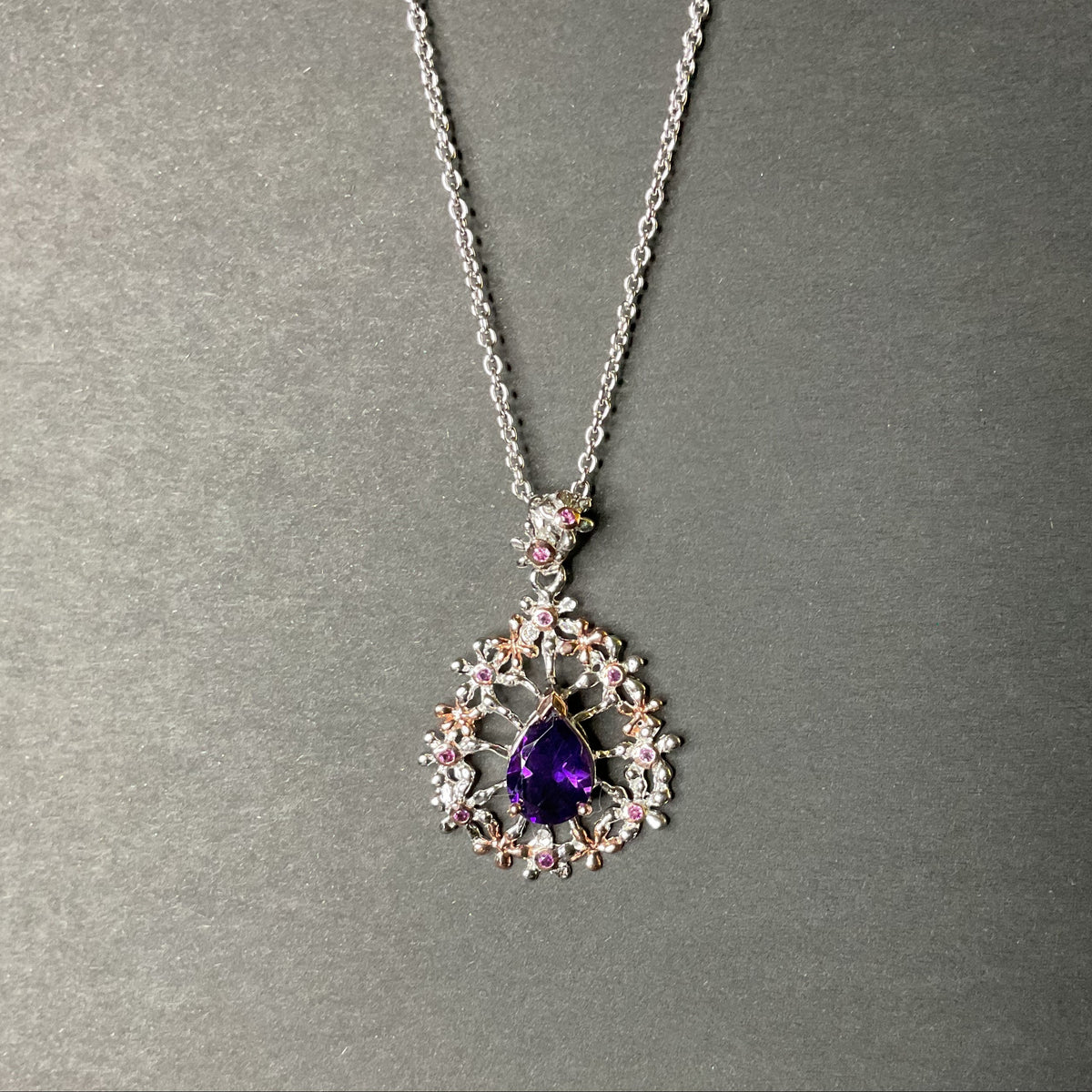 A silver pendant necklace featuring a teardrop-shaped purple gemstone surrounded by delicate floral clusters of clear and pink gemstones, displayed on a textured gray background.