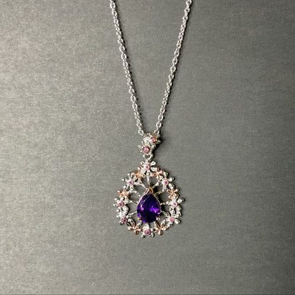 A silver pendant necklace featuring a teardrop-shaped purple gemstone surrounded by delicate floral clusters of clear and pink gemstones, displayed on a textured gray background.