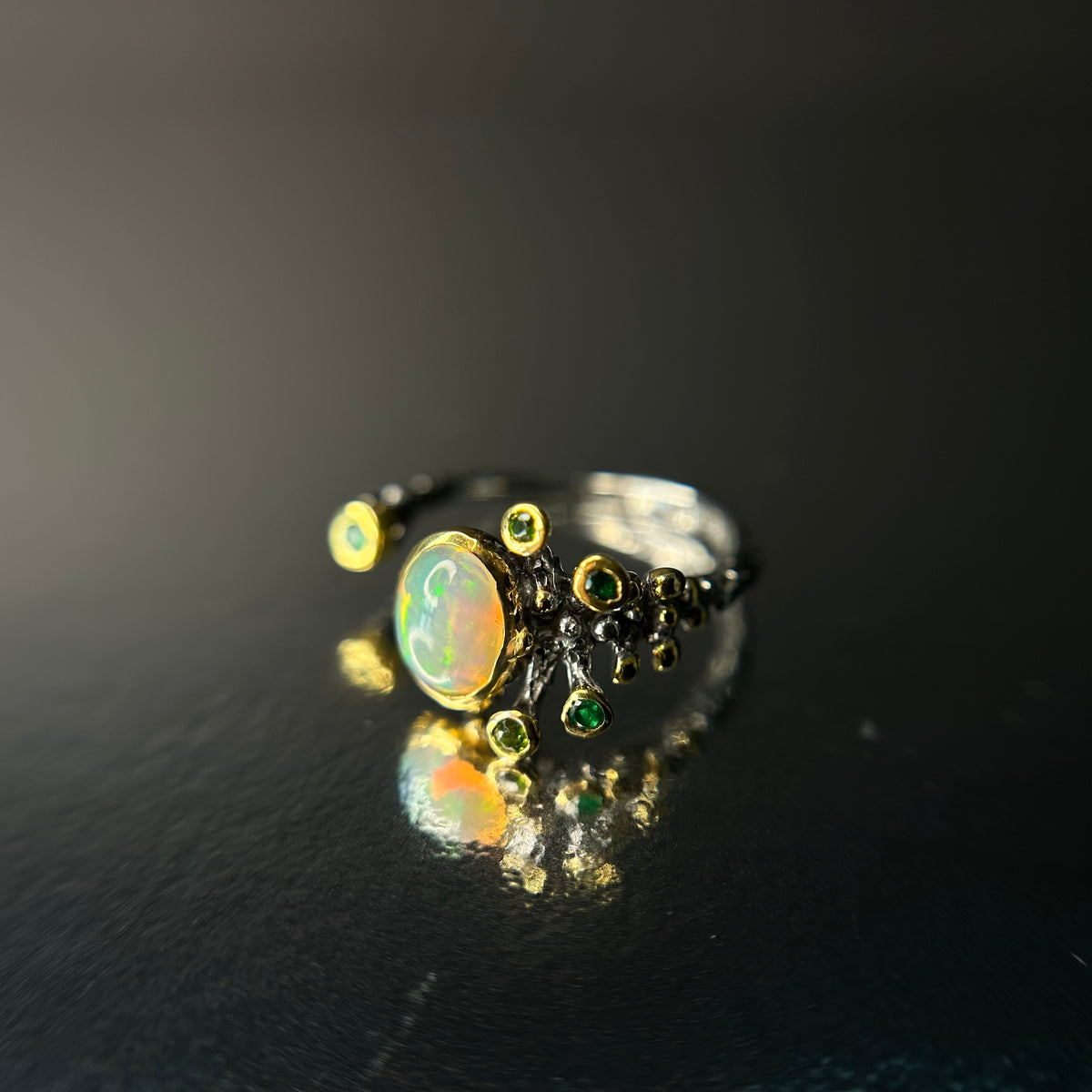 Silver ring with opal and green gemstones on a black reflective surface