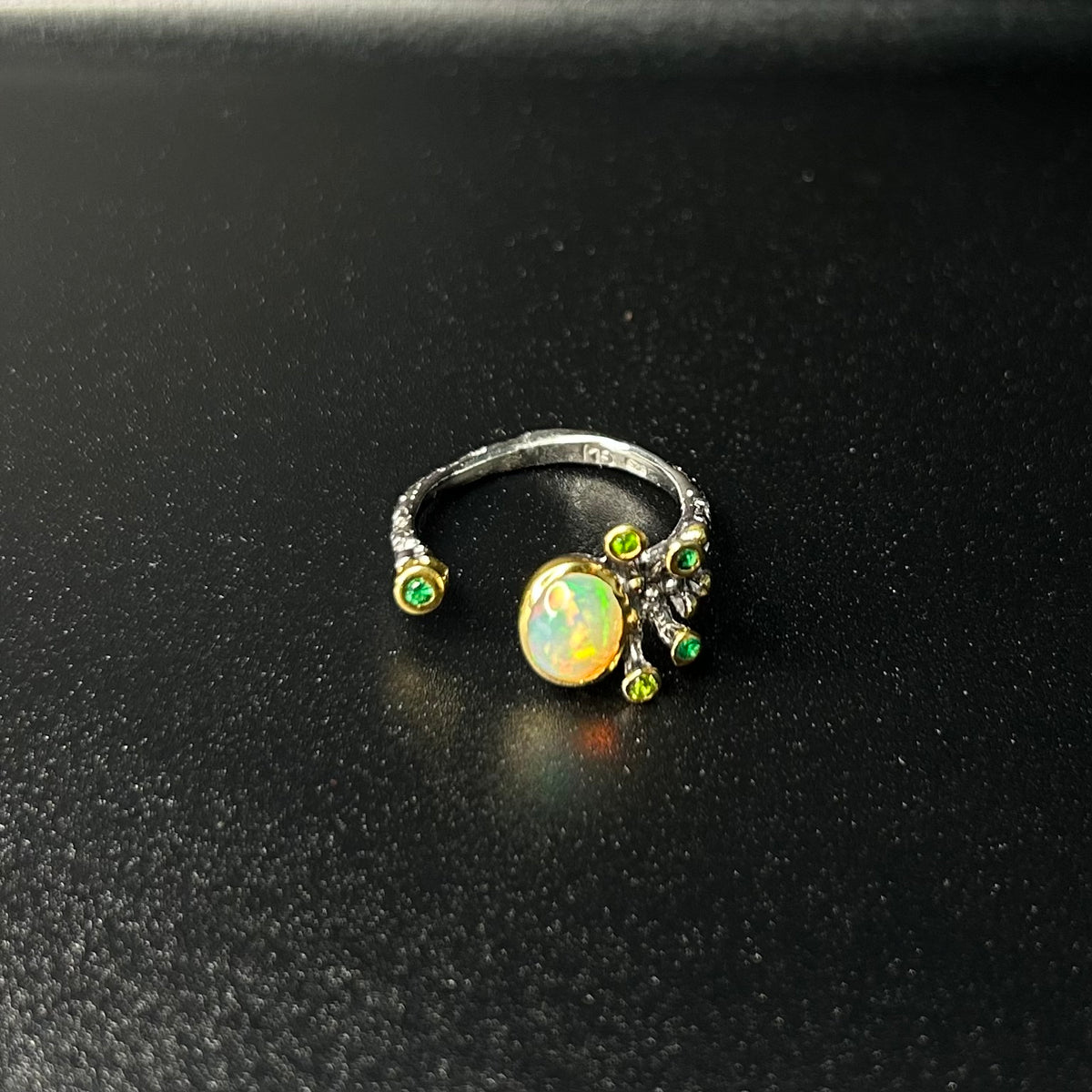 Silver ring with a central opal stone and smaller green stones on a black background