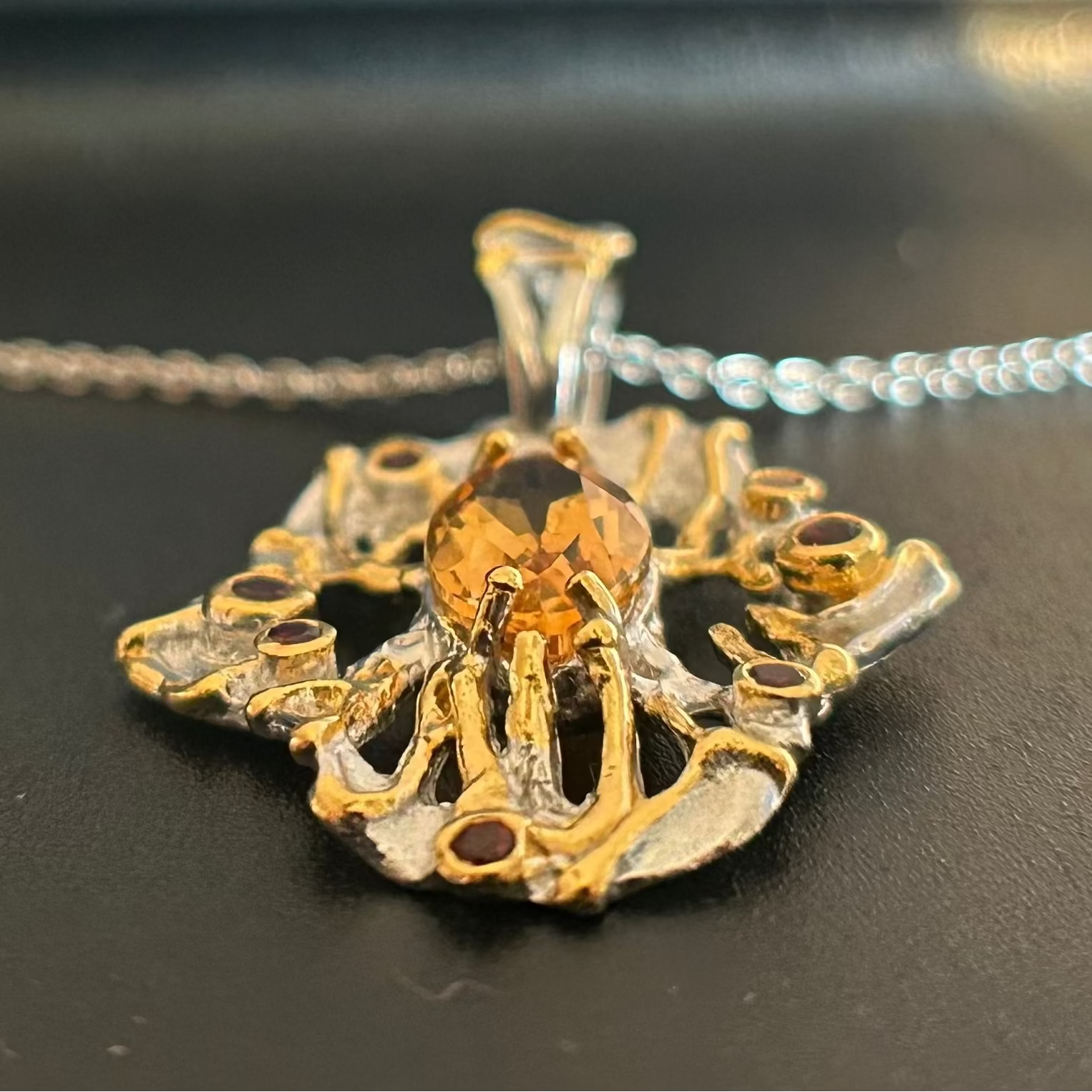 A close-up of a unique, organic-shaped pendant necklace featuring a faceted amber-orange center stone, set in a silver and gold textured metal design, with a silver chain visible in the background.