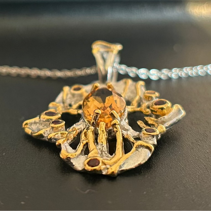 A close-up of a unique, organic-shaped pendant necklace featuring a faceted amber-orange center stone, set in a silver and gold textured metal design, with a silver chain visible in the background.