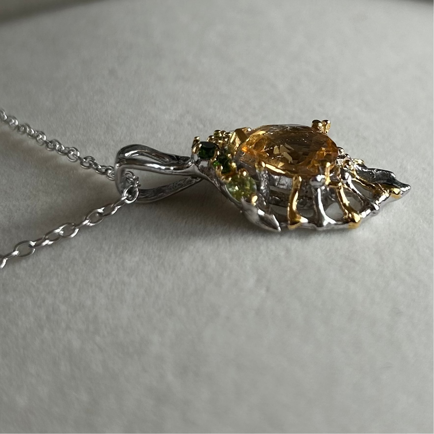 A close-up of a silver and gold pendant necklace with a central round amber-yellow Citrine gemstone and small green accent stones, resting on a light gray textured surface.
