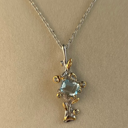 A delicate silver chain necklace with a branch-shaped pendant featuring a light blue rectangular Topaz gemstone and small clear accent Topaz stones, displayed on a neutral beige background.