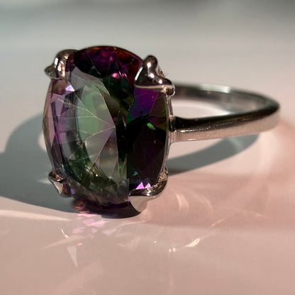 A silver ring featuring a large, faceted oval gemstone that displays a color-changing property, shifting from deep purple to green under light, set on a soft pink reflective surface.