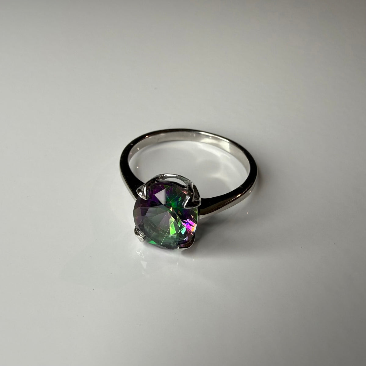 A silver ring featuring a round, faceted iridescent gemstone that shifts between green, purple, and blue, set in a four-prong setting on a plain light gray background.