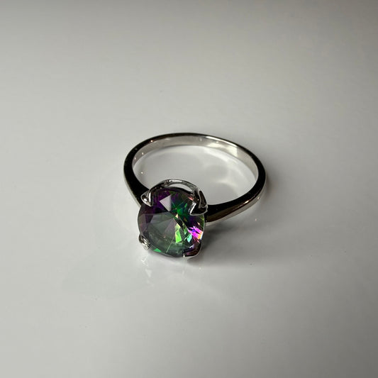 A silver ring featuring a round, faceted iridescent gemstone that shifts between green, purple, and blue, set in a four-prong setting on a plain light gray background.
