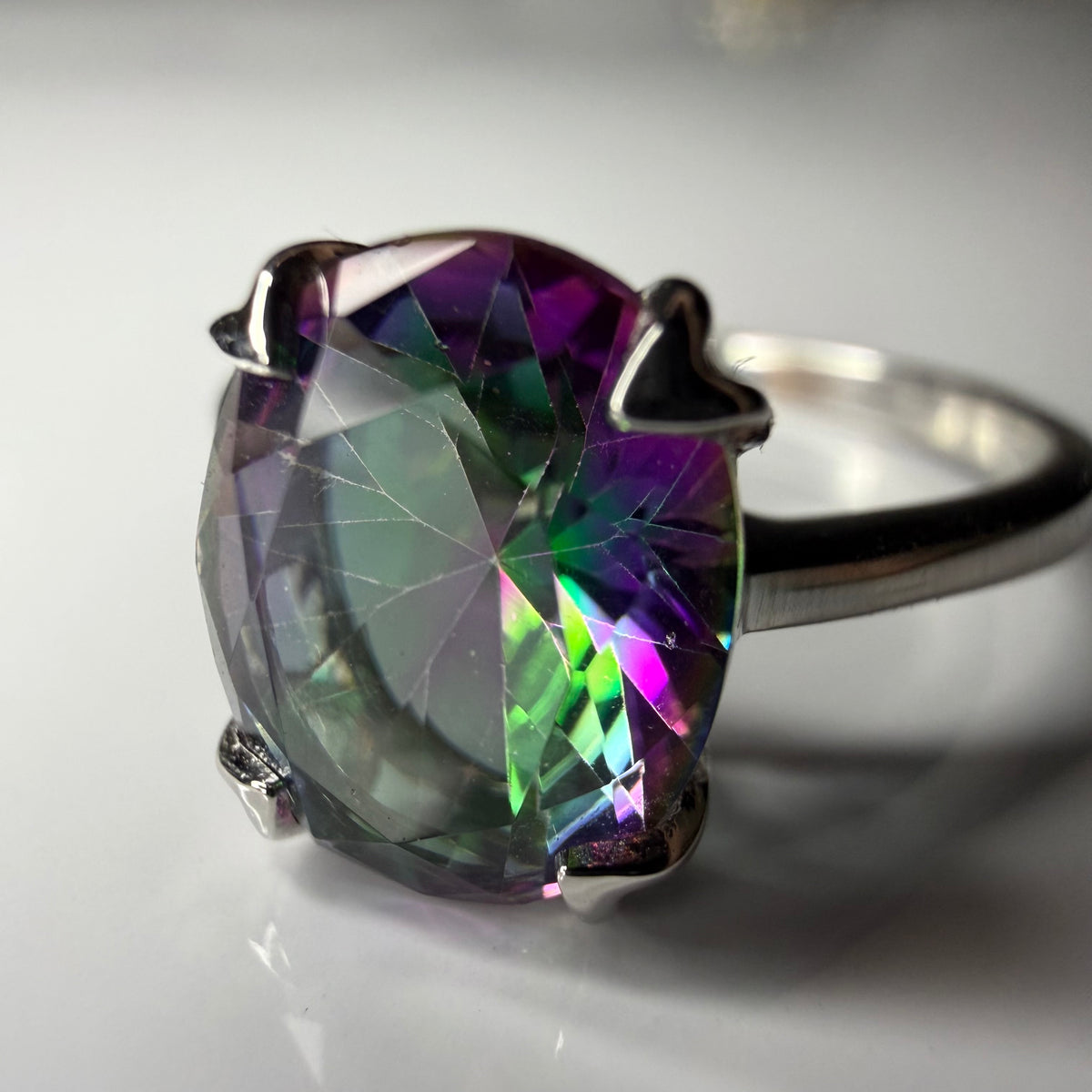 A silver ring featuring a large oval-cut iridescent gemstone that shifts between purple, green, and blue, set in a polished silver prong setting against a soft gray background.