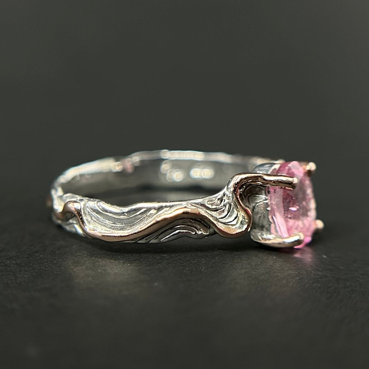 A sterling silver ring with a wavy, textured band and a cushion-cut pink gemstone, shown on a dark gray background.