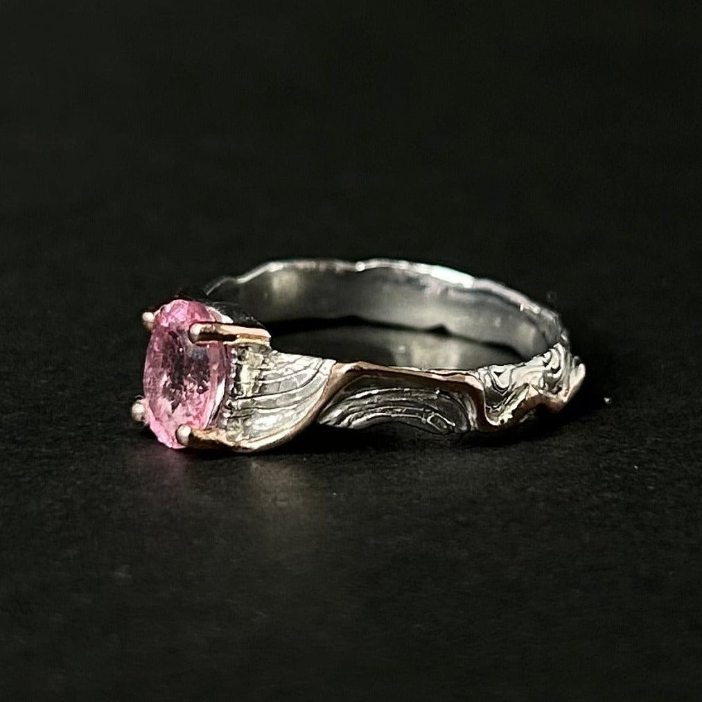 A silver ring with a wavy, textured band and a round pink tourmaline gemstone, photographed on a dark black background.