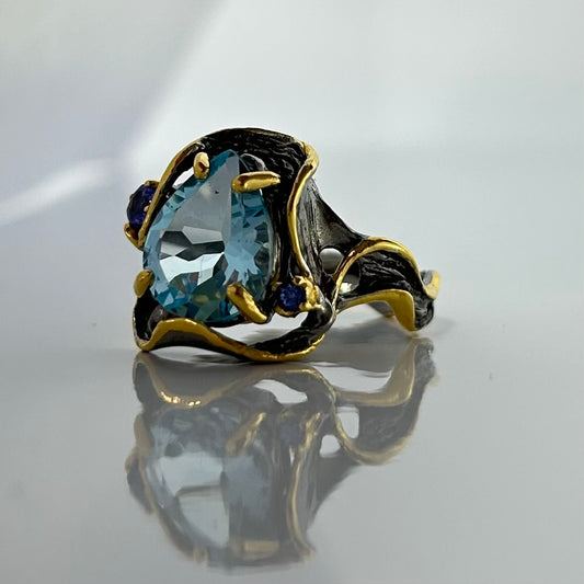 Gold and black ring with a blue topaz gemstone on a reflective surface