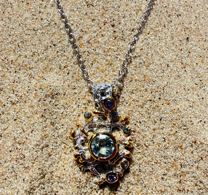 A silver necklace with a detailed, ornate pendant featuring a large round light blue Topaz gemstone and smaller dark blue and green gemstones, displayed on a sandy surface.