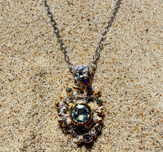 A silver necklace with a detailed, ornate pendant featuring a large round light blue Topaz gemstone and smaller dark blue and green gemstones, displayed on a sandy surface.