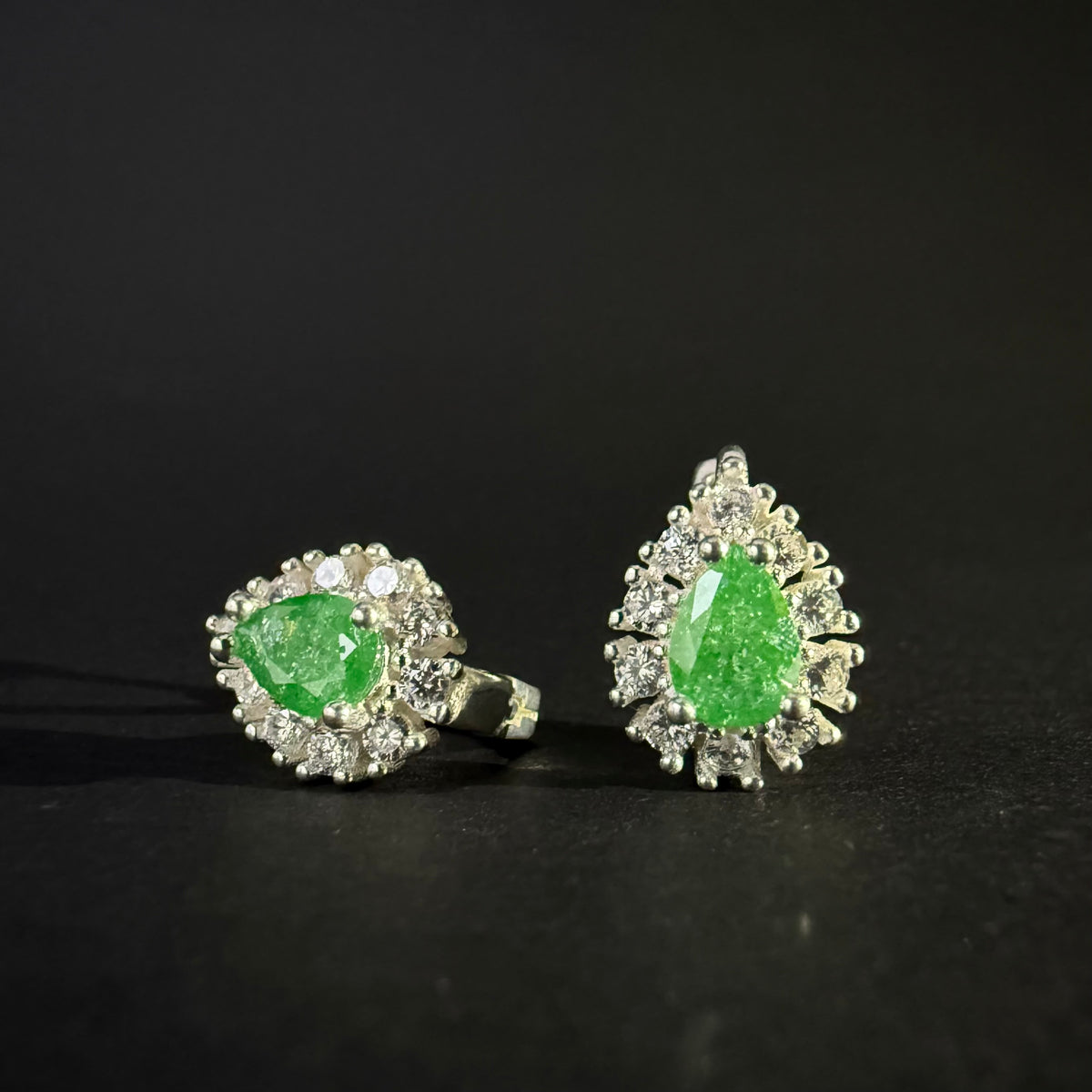 A pair of silver earrings with oval green gemstones surrounded by clear crystals, displayed on a dark reflective surface.