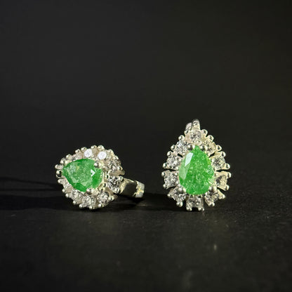 A pair of silver earrings with oval green gemstones surrounded by clear crystals, displayed on a dark reflective surface.