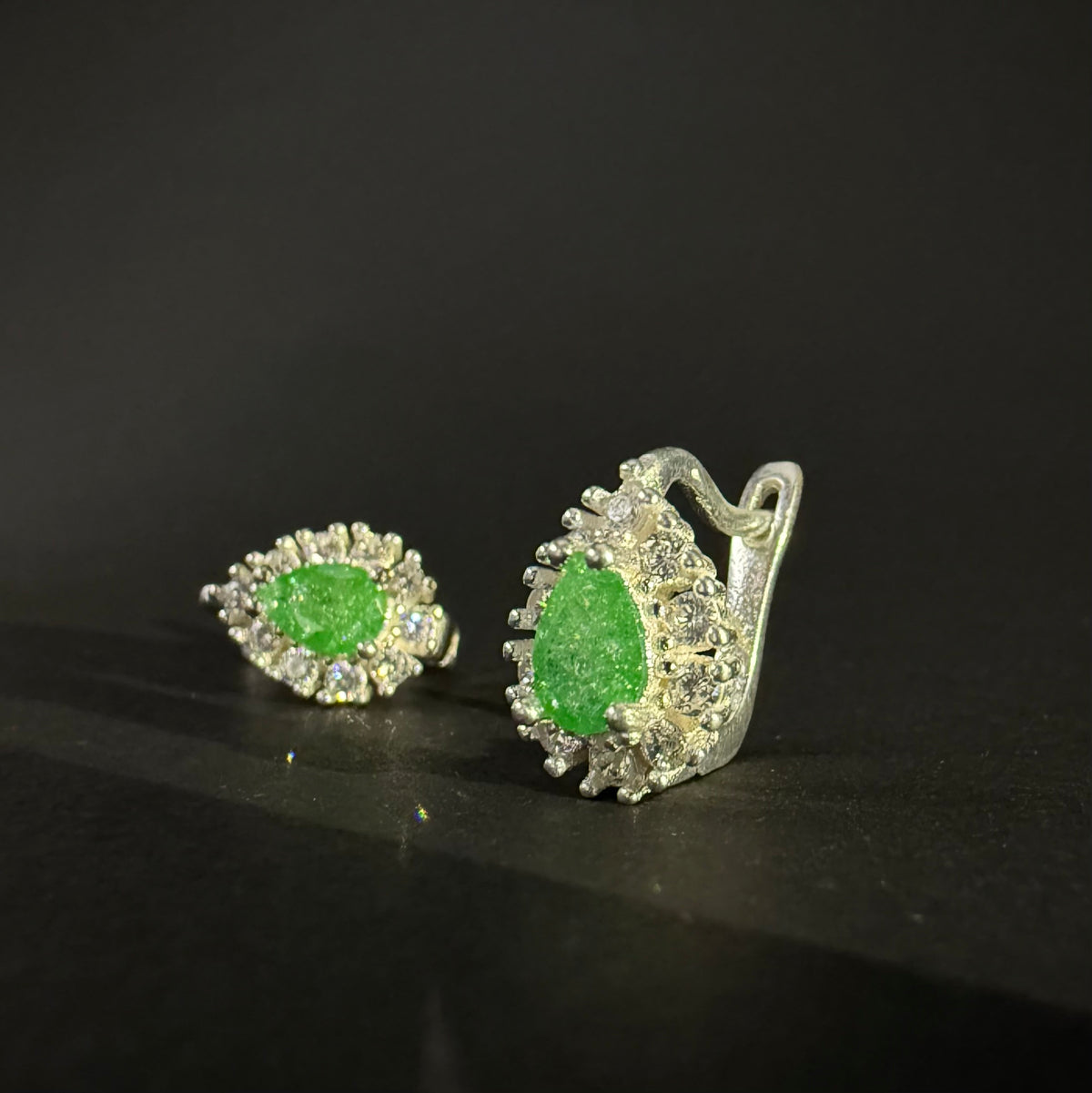 A pair of silver earrings with oval green gemstones surrounded by clear crystals, displayed on a dark reflective surface.