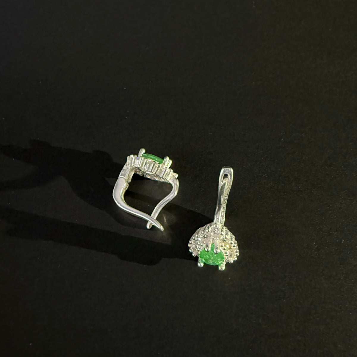 A pair of silver earrings with oval green gemstones surrounded by clear crystals, displayed on a dark reflective surface.