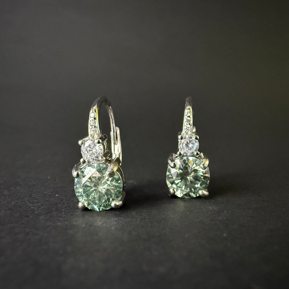 A pair of silver leverback earrings with large round-cut light blue-green moissanite and smaller clear stones.