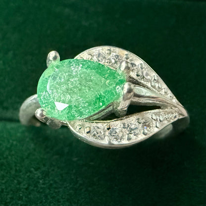 A silver ring featuring a large, faceted green gemstone in an oval shape, surrounded by small clear stones set in a leaf-like design