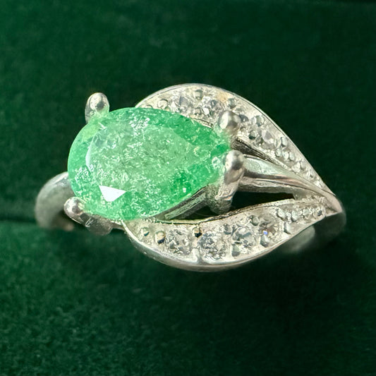 A silver ring featuring a large, faceted green gemstone in an oval shape, surrounded by small clear stones set in a leaf-like design