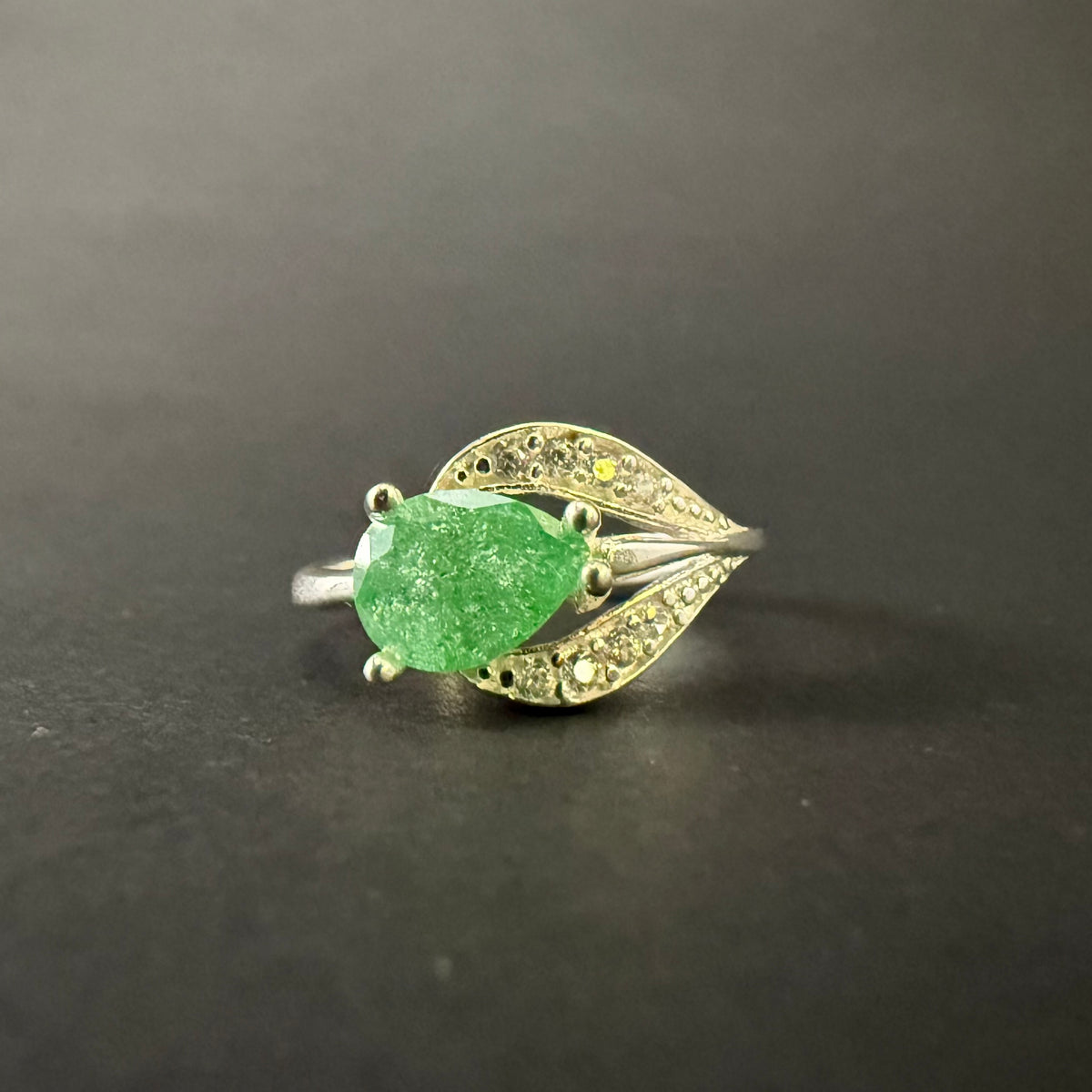A silver ring featuring a large, faceted green gemstone in an oval shape, surrounded by small clear stones set in a leaf-like design