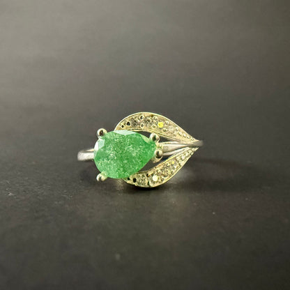 A silver ring featuring a large, faceted green gemstone in an oval shape, surrounded by small clear stones set in a leaf-like design