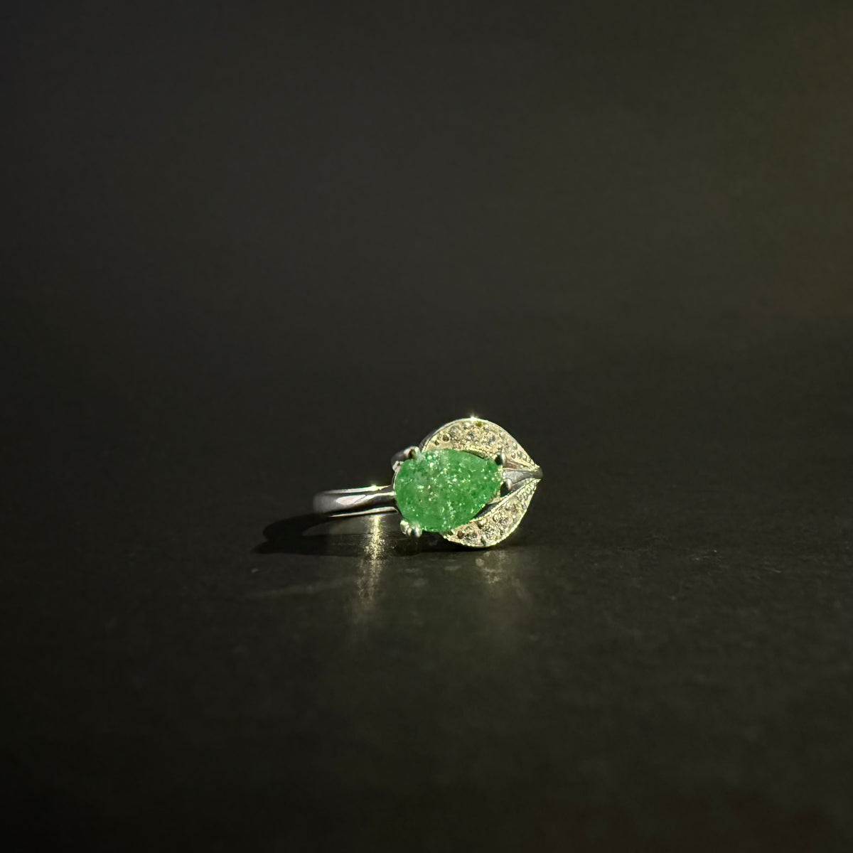A silver ring featuring a large, faceted green gemstone in an oval shape, surrounded by small clear stones set in a leaf-like design