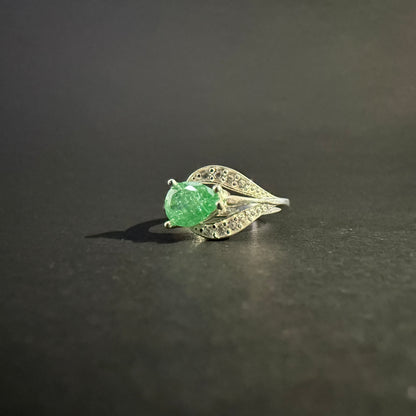 A silver ring featuring a large, faceted green gemstone in an oval shape, surrounded by small clear stones set in a leaf-like design
